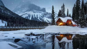 雪中小屋的溫暖燈光映照在群山環繞的冰凍湖面上。 — 冬天 壁紙