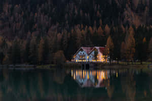 舒適的高山小屋在黃昏時發光，在寧靜的湖面上完美倒映。 — MacBook Air 15 壁紙