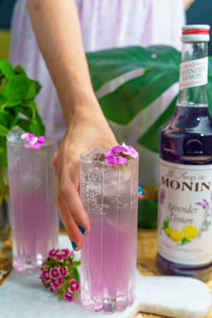 手拿著薰衣草檸檬飲料，配以粉紅花朵和Monin糖漿瓶。 — Vivo Pad 壁紙