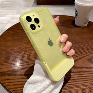 手拿著帶有心形貼紙的黃色手機殼，放在木桌上，靠近咖啡杯。 — 美學 iPhone 壁紙