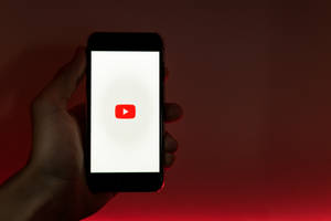 手拿著手機，在紅色背景上顯示 YouTube 應用程式圖標 — 手機 壁紙