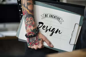 有紋身的手拿著寫有「Be Creative Design」文字的記事板。 — 黃色美學 壁紙