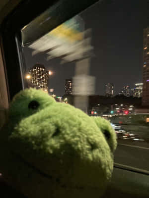 綠色毛絨青蛙在夜晚從火車窗戶向外看，城市燈光 — Kindle 壁紙