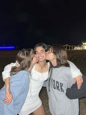 三個年輕女性在夜間海灘上擁抱並親吻臉頰，伴有藍色燈光。 — 老友記 壁紙