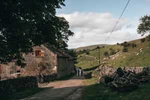 三個人走在鄉間泥路上，靠近一座石屋，背景是綠色山丘。 — 鄉村風 壁紙