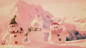 三個戴帽和圍巾的雪人裝飾品，站在雪地上，背景有山。 — 冬日美學 壁紙