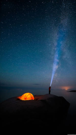 夜晚在懸崖上露營的人，發光的帳篷和指向銀河的手電筒。