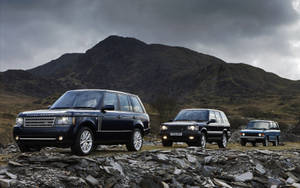 三輛 Range Rover SUV 停放在岩石地面上，背景有山脈，天空多雲。