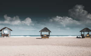 三間茅草海灘小屋在沙灘上，有海浪和烏雲天空。 — 2880x1800 壁紙