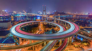 夜間發光的螺旋形高速公路，車燈流光，背景是城市天際線。 — 中國 壁紙