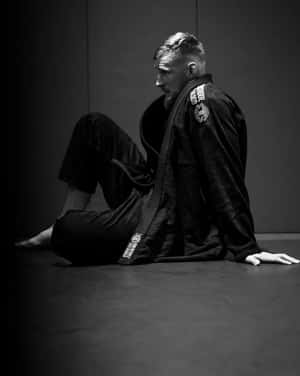 一個專注的武術家穿著黑色道服，坐在黑暗練習室的訓練墊上。 — 黑白 壁紙