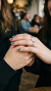 兩隻手，帶有彩虹指甲和銀戒指，在咖啡廳中交握。 — 女同志 GIF 壁紙