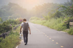一位旅行者背著背包和滑板車走在風景如畫的鄉村道路上。 — 夏日氣息 壁紙