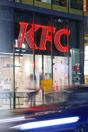 一個發光的紅色KFC標誌在玻璃外牆的餐廳中，外面有模糊的人影和汽車移動。 — 小米Pad 7 Ultra 壁紙