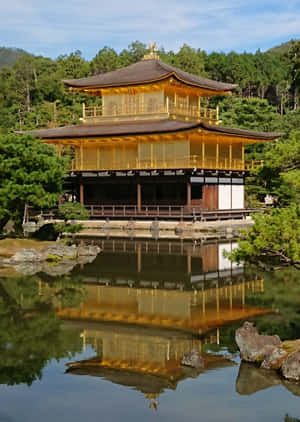 金色寺廟倒映在平靜池塘中，伴隨著綠樹和藍天 — 日本 壁紙