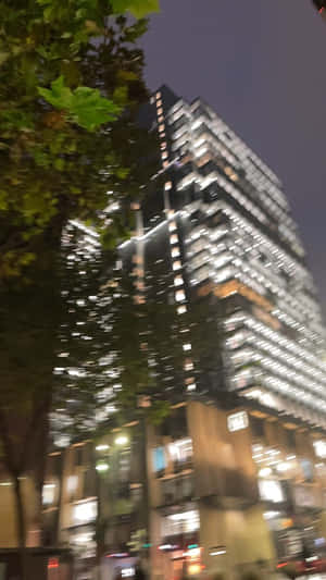 夜晚亮燈的高聳玻璃建築，前景有模糊的樹木。