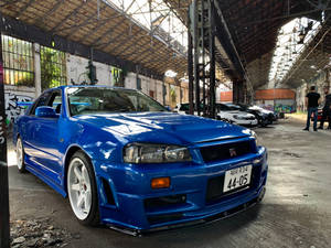 藍色日產Skyline R34停放在一個佈滿塗鴉的倉庫中，背景有人群。 — 藍色 壁紙