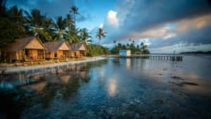 熱帶海灘茅草屋頂小屋在平靜的碧綠海水中倒映，背景是戲劇性的黃昏天空。