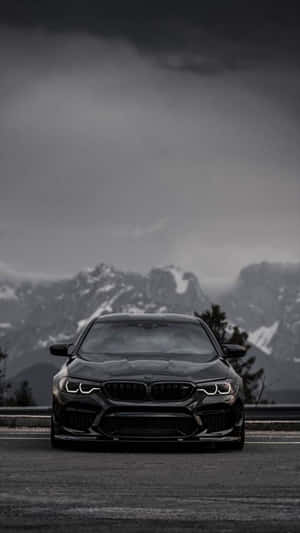黑色BMW M5停在山路上，背景有白雪覆蓋的山峰和風暴般的天空。