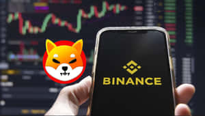 手握住手機，帶有 Binance 標誌和 Shiba Inu 圖標，背景為股票圖表。