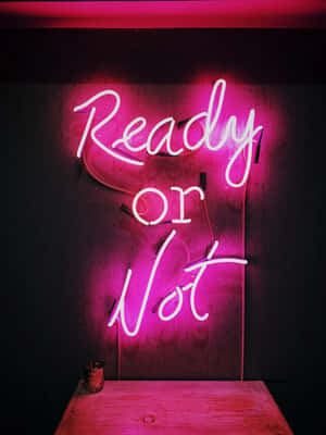 粉紅色霓虹燈牌，上面寫著「Ready or Not」，位於黑暗的牆壁上，下方有一張木桌。