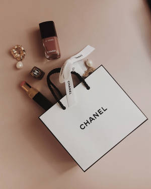 Chanel 購物袋搭配指甲油、唇膏和珠寶，置於粉紅色表面上。