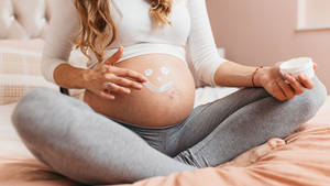 孕婦坐在床上，在肚子上塗抹乳液形成笑臉形狀。