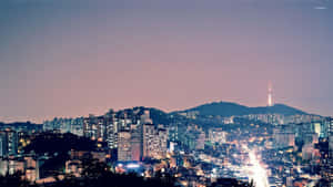 黃昏時分的城市天際線，建築物燈火通明，山丘上有一座塔樓。