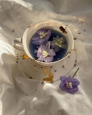 一杯藍色茶，帶有漂浮的紫色花朵，放在白色布料上，散落著花瓣。 — 4:5 壁紙