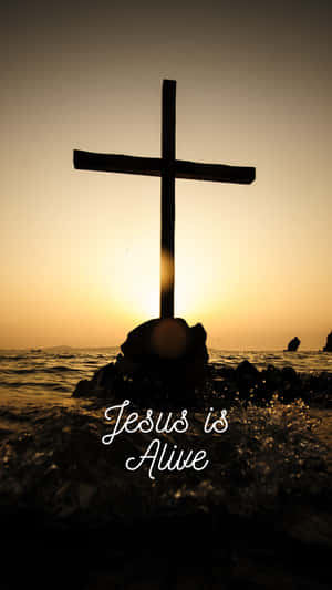 日落時岩石上十字架的剪影，伴隨海浪和文字 Jesus is Alive