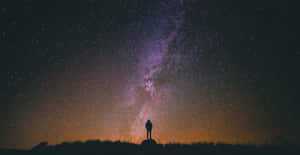 一個人在山丘上的剪影，背景是充滿銀河系的鮮豔星空。 — 月色夜空 壁紙