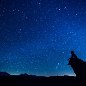 人坐在懸崖邊，星空下，背景有山脈 — 太空 壁紙
