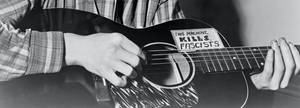 雙手彈奏結他，結他上有寫著「This Machine Kills Fascists」的政治貼紙。 — 黑白 壁紙