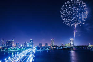 煙花在夜間城市天際線上空綻放，燈光倒映在水面上。