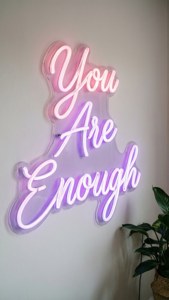 粉紅色和紫色的霓虹燈牌，寫著 You Are Enough，附近有一棵植物。