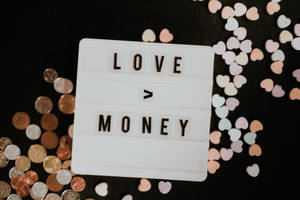 白色燈箱上寫著「LOVE > MONEY」，周圍有硬幣和粉彩心形，黑色背景。