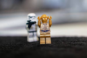 金色C-3PO LEGO人偶，舉起手臂，後面有模糊的風暴兵，黑色表面。