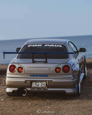 銀色日產Skyline R34藍色尾翼停泊在岩石海岸近海。