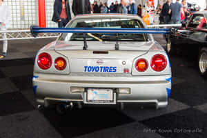 銀色日產Skyline R34，配備藍色尾翼，於室內車展中。