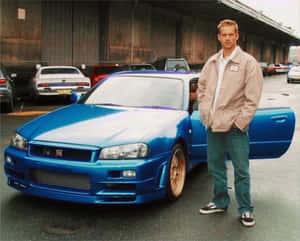 身穿米色外套的男子站在天橋下的藍色日產Skyline R34旁邊。
