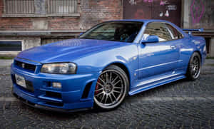 藍色日產Skyline R34跑車停在鵝卵石街道上，靠近塗鴉牆。