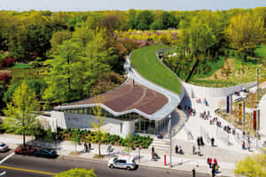 現代建築，玻璃牆和弧形屋頂，位於綠色公園中，有人們漫步。 — MacBook Air M2 壁紙