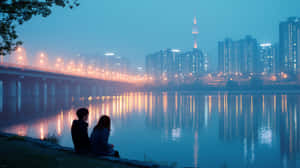 夜晚，兩個人坐在河邊，城市燈光倒映在水中。 — 夜晚 GIF 壁紙