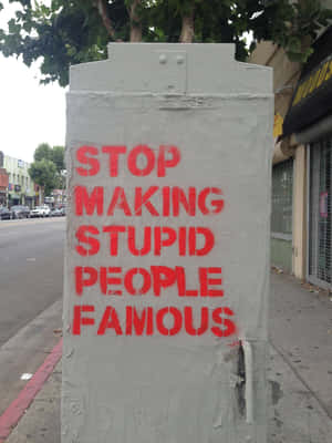 灰色公用箱上嘅紅色塗鴉寫住「STOP MAKING STUPID PEOPLE FAMOUS」，喺城市行人路上。