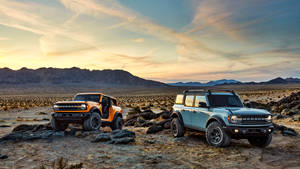 夕陽下停在岩石沙漠中的兩輛Bronco，背景有山脈。 — 老福特貨車 壁紙