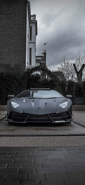 一輛灰色的林寶堅尼 Aventador 停泊在濕漉漉的鵝卵石街道上，天空多雲。