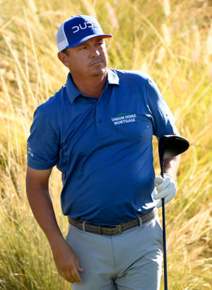 穿藍色襯衫和帽子的高爾夫球手在陽光明媚的日子裡在高高的草叢中握著球桿 — 運動 壁紙