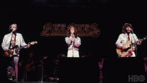 Bee Gees 身穿白色服裝，手持結他和咪高峰在舞台上現場表演。 — 荷里活 GIF 壁紙