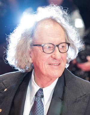 一位白髮戴眼鏡的年長男性在正式西裝中於活動中微笑。