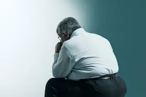 一位身穿白襯衫的年長男性,靠著藍白牆壁,低著頭,以沉思的姿勢彎腰坐著。